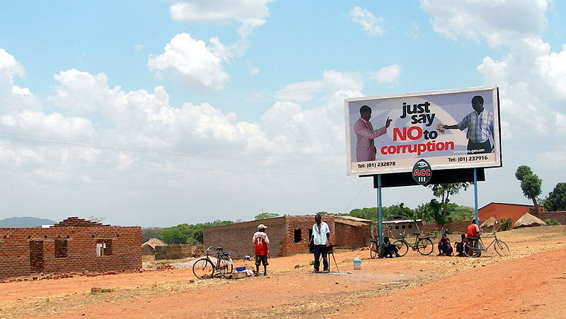 A billboard with the message "just say NO to corruption" featuring two men shaking hands, set against a rural landscape with brick houses, people with bicycles, and a partly cloudy sky.