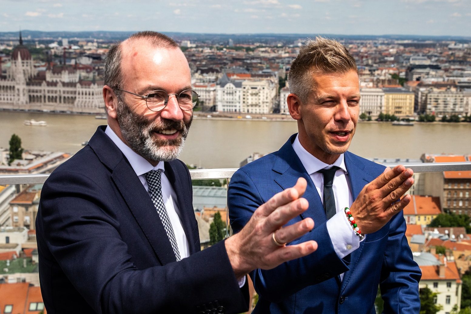 Péter Magyar and Manfred Weber standing side by side with a cityscape featuring the Hungarian Parliament building and the Danube River in the background.
